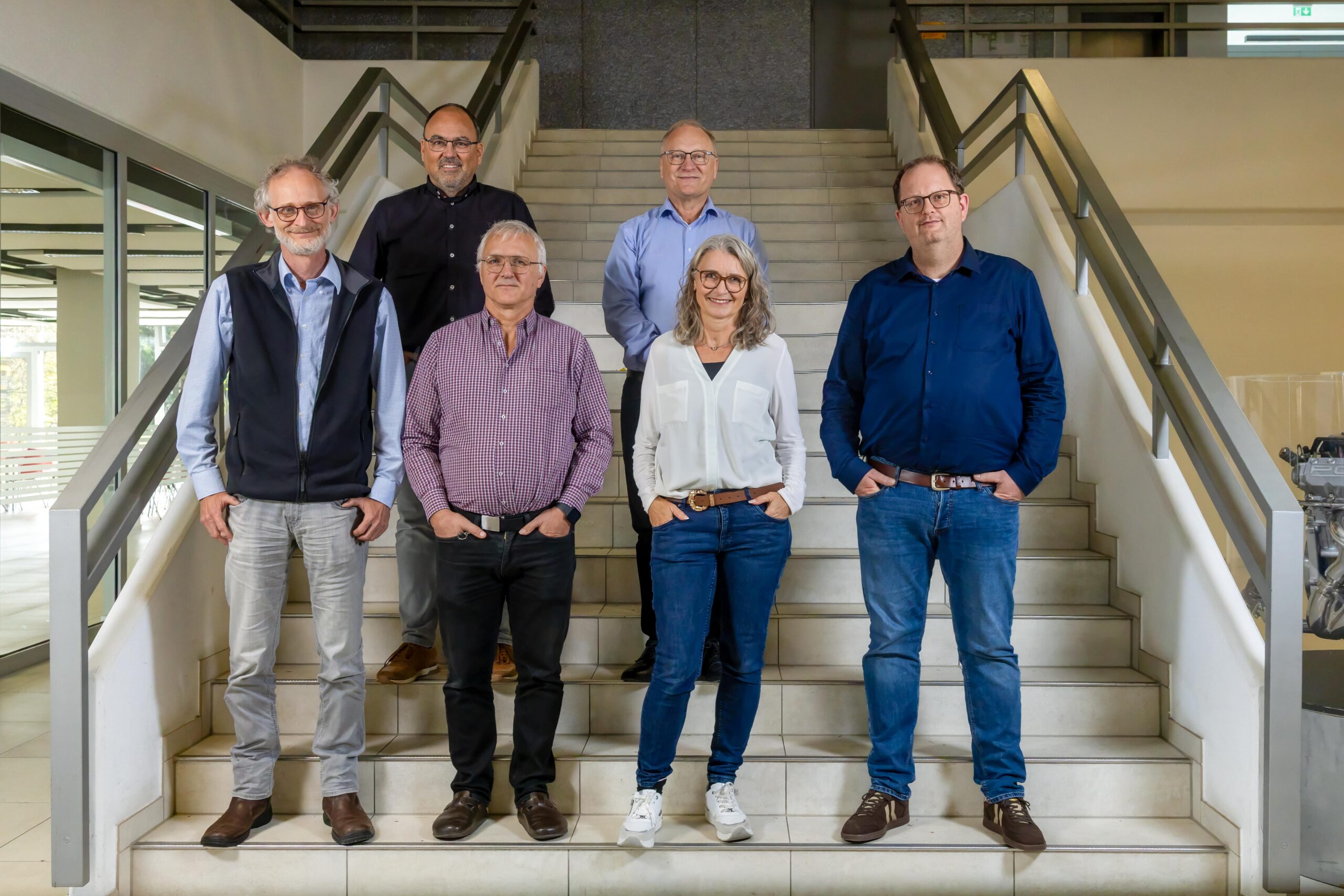 Die sechs Mitglieder des Schulleitungsteams  stehen auf einer breiten Treppe bei der Aula im Schulgebäude Villingen. Sie tragen überwiegend Freizeitkleidung wie Hemden, Jeans und Pullover. Die Hände sind meist in den Hosentaschen. Im Hintergrund sind Glasflächen und helle Wände zu sehen. Die Szene wirkt sachlich und neutral.